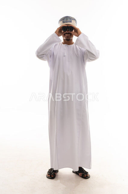 Looking and smiling in front of the camera, an Arab Gulf Omani man wearing a white dishdasha and turban holding binoculars and looking at something, gestures of happiness and joy, full-body, portrait with a white background.