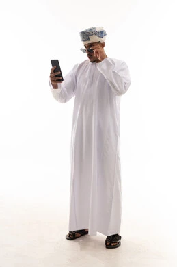 رجل عربي خليجي عماني يرتدي الدشداشة البيضاء و العمامة يقوم بنزع النظارة الشمسية اثناء استخدام الهاتف المحمول لرؤية اوضح , الجسم بالطول الكامل , بورتريه خلفية بيضاء