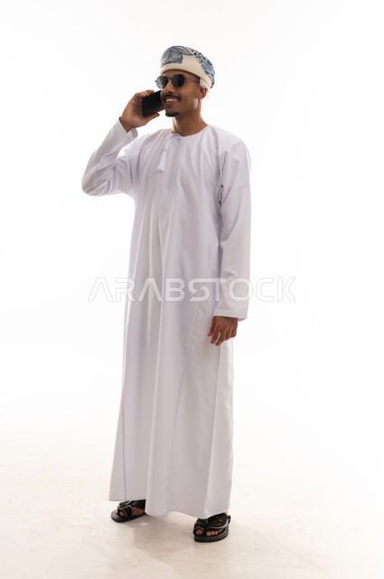 رجل عربي خليجي عماني يرتدي الدشداشة البيضاء و العمامة يقوم بالتحدث في الهاتف المحمول , ايماءات السعادة اثناء اجراء المكالمات الصوتية , الجسم بالطول الكامل , بورتريه خلفية بيضاء