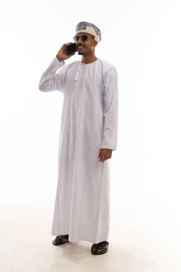 رجل عربي خليجي عماني يرتدي الدشداشة البيضاء و العمامة يقوم بالتحدث في الهاتف المحمول , ايماءات السعادة اثناء اجراء المكالمات الصوتية , الجسم بالطول الكامل , بورتريه خلفية بيضاء