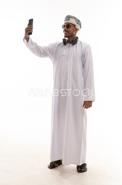 Taking a selfie and recording special moments, an Arab Gulf Omani man wearing a white dishdasha and a turban holding a mobile phone, gestures of joy and happiness, smiling at the phone, portrait with a white background, full body.