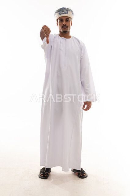 رجل عربي خليجي عماني يرتدي الدشداشة البيضاء و العمامة يرفع اصبع الابهام لاسفل علامة على عدم الاعجاب , ايماءات الانزعاج من شئ ما , بورتريه خلفية بيضاء , الجسم بالطول الكامل