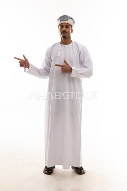 رجل عربي خليجي عماني يرتدي الدشداشة البيضاء و العمامةيرفع اصبعي السبابة للجانب يشير الي شئ ما , نظرة امامية للكاميرا , بورتريه خلفية بيضاء , الجسم بالطول الكامل