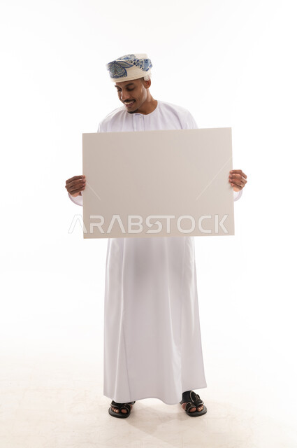 رجل عربي خليجي عماني يرتدي دشداشة بيضاء و عمامة يحمل ورقة كبيرة و ينظر للاسفل, تعابير السرور و الفرحة , الوقوف امام الكاميرا , الجسم بالطول الكامل , بورتريه خلفية بيضاء