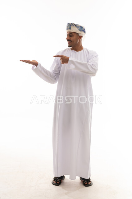 Looking to the side while pointing with the index finger, an Arab Gulf Omani man wearing a turban and a white dishdasha stands in front of the camera, with expressions of happiness and joy, portrait with a white background, full body.