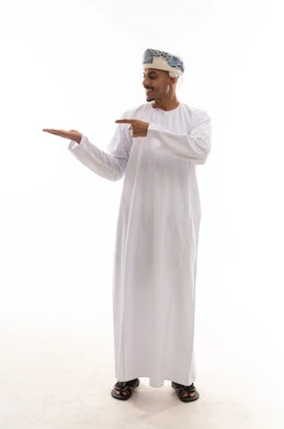 Looking to the side while pointing with the index finger, an Arab Gulf Omani man wearing a turban and a white dishdasha stands in front of the camera, with expressions of happiness and joy, portrait with a white background, full body.