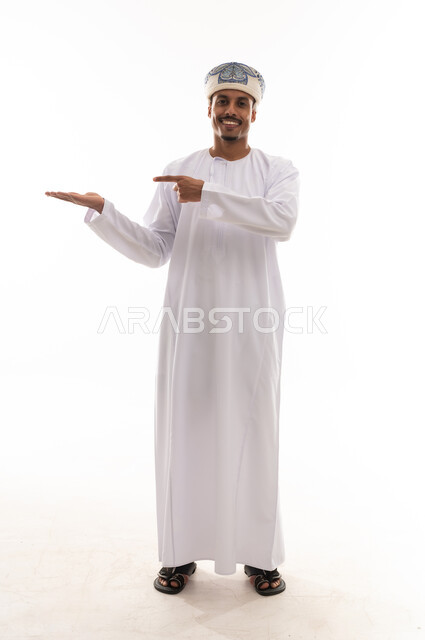 الاشارة باصبع السبابة الي شئ ما و فتح قبضة اليد الاخري, رجل عربي خليجي عماني يرتدي دشداشة بيضاء و عمامة , الوقوف و النظر  امام الكاميرا , بورتريه خلفية بيضاء , الجسم بالطول الكامل ,