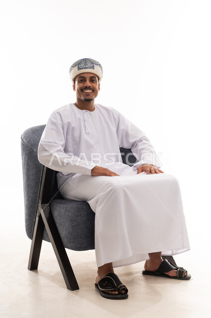 نظرة أمامية للكاميرا مع الابتسامة , رجل عربي خليجي عماني يرتدي عمامة و دشداشة بيضاء , الجلوس مع تعابير الفرحة و البهجة , الجسم بالطول الكامل , بورتريه الجسم بالطول الكامل