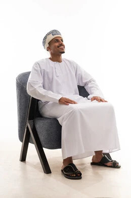 النظر بسعادة و ابتسامة للاعلى الى شئ ما , رجل عربي خليجي عماني يرتدي دشداشة بيضاء و عمامة يضع اليدين على رجله  اثناء الجلوس , تعابير الفرحة و السررور , الجسم بالطول الكامل , خلفية بيضاء