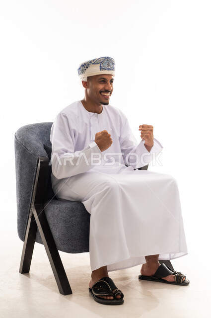 Clenching his fists tightly and raising them up is a sign of immense happiness and joy. A cheerful Arab Gulf Saudi man wearing a white thobe and a headscarf sits on a gray chair, portrait with a white background, full-body.