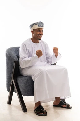 قفل قبضة اليدين باحكام و رفعها للاعلى اشارة الي السعادة و الفرحة العارمة , رجل عربي خليجي سعودي مبتهج يرتدي دشداشة بيضاء و عمامة  يجلس على كرس رمادي , بورتريه خلفية بيضاء , الجسم بالطول الكامل