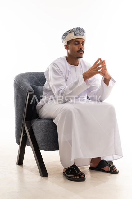 The movement of intertwining fingers is an expression of tension and anxiety. An Arab Gulf Omani man wearing a white dishdasha and a turban sits on a gray chair, looking at the intertwined fingers. Portrait with a white background, full-body.
