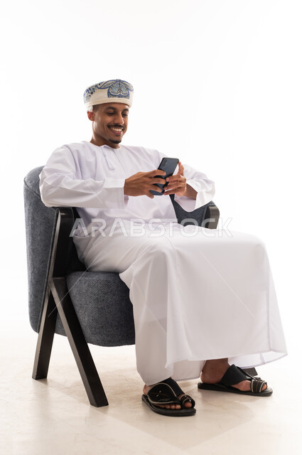 النظر و الابتسام الى شاشة الهاتف المحمول , رجل عربي خليجي عماني يرتدي العمامة و دشداشة بيضاء يجلس على كرسي رمادي و يشعر بالسعادة  , بورتريه خلفية بيضاء , الجسم بالطول الكامل