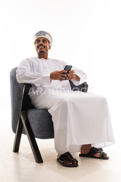 رجل عربي خليجي عماني يرتدي عمامة ودشداشة بيضاء يحمل الهاتف المحمول و يجلس على كرسي رمادي اللون , النظر الى شئ ما , بورتريه خلفية بيضاء , ايماءات السرور والفرحة , الجسم بالطول الكامل