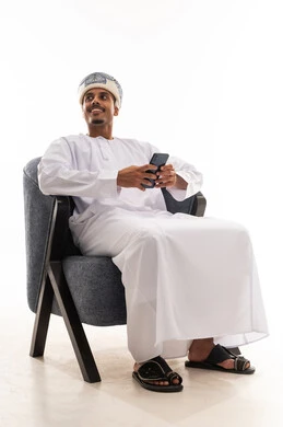 An Arab Gulf Omani man wearing a turban and a white dishdasha is holding a mobile phone and sitting on a gray chair, looking at something, portrait with a white background, gestures of joy and happiness, full body.