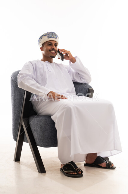 Making voice calls easily with multiple applications, an Arab Gulf Omani man wearing a white dishdasha and a turban is talking on a mobile phone, gestures of happiness and smiling, portrait with a white background, full body.