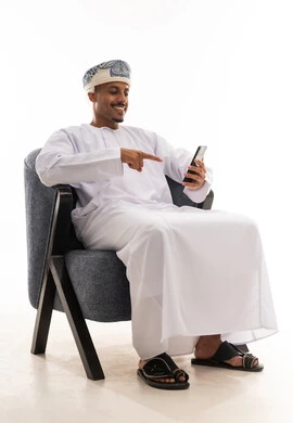 سهولة التواصل و الاتصال بالاهل و الاصدقاء , رجل عربي خليجي عماني يرتدي دشداشة بيضاء و عمامة يشير باصبع السبابة الي شاشة الهاتف المحمول اثناء محادثة الفيديو , ايماءات السعادة و الابتسام الي شاشة الجوال , بورتريه خلفية بيضاء , الجسم بالطول الكامل