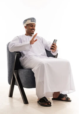 اجراء مكالمات الفيديو المباشرة و البقاء متصل مع العائلة و الاصدقاء , رجل عربي خليجي عماني يرتدي دشداشة بيضاء و عمامة يتواصل مع الاهل باستخدام التطبيقات الحديثة المتعددة , بورتريه خلفية بيضاء , الجسم بالطول الكامل