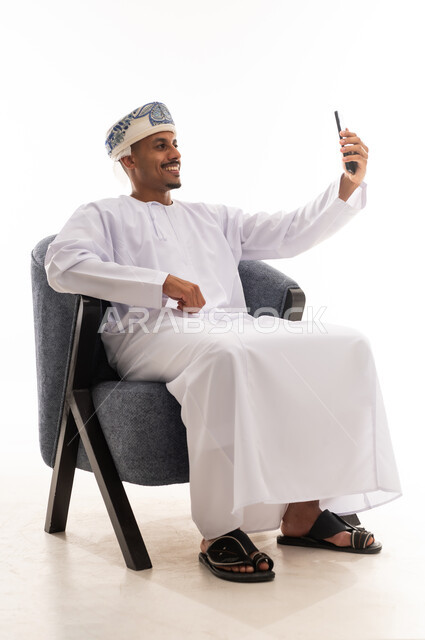 التقاط الصور الشخصية في سعادة , رجل عربي خليجي عماني يرتدي دشداة بيضاء و العمامة يرفع الهاتف المحمول و يلتقط صورة فوتوغرافية لنفسه , مفهوم التطور التقني  , بورتريه خلفية بيضاء , الجسم بالطول الكامل