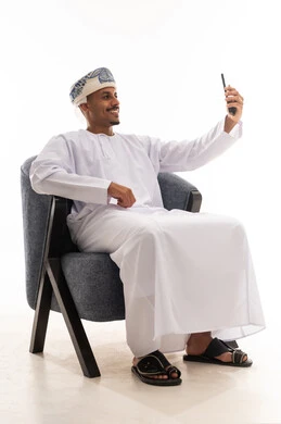 التقاط الصور الشخصية في سعادة , رجل عربي خليجي عماني يرتدي دشداة بيضاء و العمامة يرفع الهاتف المحمول و يلتقط صورة فوتوغرافية لنفسه , مفهوم التطور التقني  , بورتريه خلفية بيضاء , الجسم بالطول الكامل