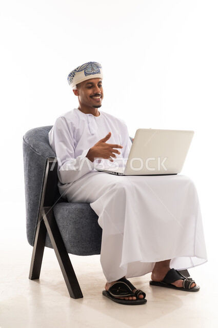 التلويح و تحريك اليدين اثناء الحديث المباشر باستخدام التطبيقات المتطورة , تعابير الحماس و الاستمتاع , رجل عربي خليجي عماني يرتدي دشداشة بيضاء و عمامة يحمل الحاسوب المتنقل بسهولة , بورتريه خلفية بيضاء , الجسم بالطول الكامل