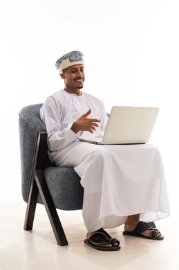 التلويح و تحريك اليدين اثناء الحديث المباشر باستخدام التطبيقات المتطورة , تعابير الحماس و الاستمتاع , رجل عربي خليجي عماني يرتدي دشداشة بيضاء و عمامة يحمل الحاسوب المتنقل بسهولة , بورتريه خلفية بيضاء , الجسم بالطول الكامل