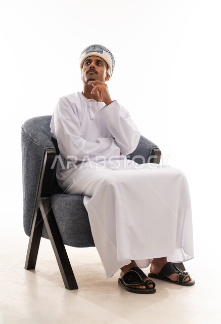 رجل عربي خليجي عماني يرتدي دشداشة بيضاء و عمامة يضع يده على وجهه اشارة الي التفكير العميق و الحيرة , النظر للجانب الى شئ ما , بورتريه خلفية بيضاء , الجسم بالطول الكامل