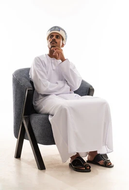 رجل عربي خليجي عماني يرتدي دشداشة بيضاء و عمامة يضع يده على وجهه اشارة الي التفكير العميق و الحيرة , النظر للجانب الى شئ ما , بورتريه خلفية بيضاء , الجسم بالطول الكامل