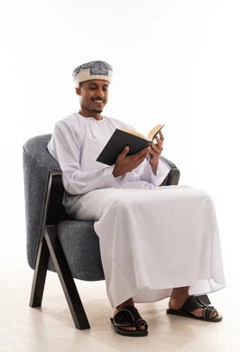 قراءة الكتب المسلية المضحكة , رجل عربي خليجي عماني يرتدي الثوب الابيض التقليدي في سلطنة عمان و عمامة ينظر الى الكتاب بابتسامة , ايماءات السعادة و الفرحة , بورتريه خلفية بيضاء , الجسم بالطول الكامل