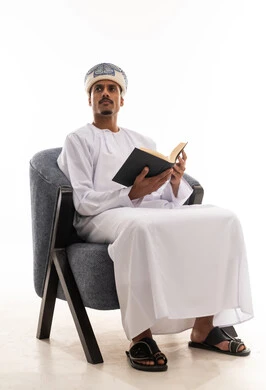 Looking to the side at something, an Arab Gulf Omani man wearing a white dishdasha and a turban reading an interesting book, immersed in deep thought, full-body portrait with a white background.