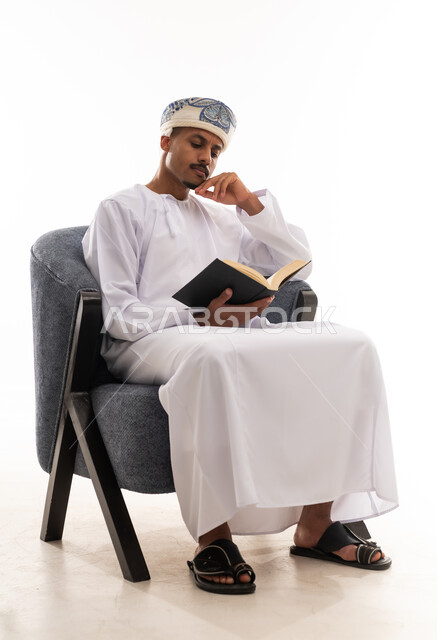 بورتريه خلفية بيضاء , الجسم بالطول الكامل , رجل عربي عماني خليجي يرتدي دشداشة بيضاء و عمامة يضع يده على الوجه اشارة الي التركيز والاستغراق في القراءة الممتعة ,