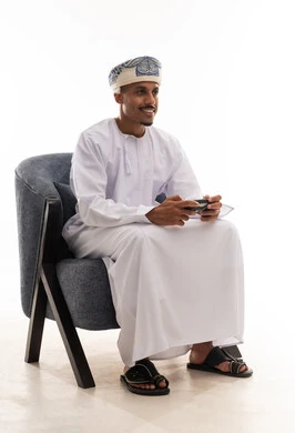 قضاء الاوقات المسلية , ممارسة الانشطة الترفيهية , رجل عربي خليجي عماني يرتدي دشداشة بيضاء و عمامة يلعب العاب فيديو الكترونية منزلية , ايماءات الفرحة و السرور , الجسم بالطول الكامل , بورتريه خلفية بيضاء