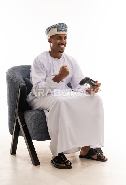 قفل فبضة اليد باحكام والتلويح بعلامة النصر و تحقيق الفوز , رجل عربي خليجي عماني يرتدي دداشة بيضاء و عمامة يحمل يد التحكم بلعبة الفيديو الالكترونية , ايماءات السعادة و البهجة , بورتريه خلفية بيضاء , الجسم بالطول الكامل