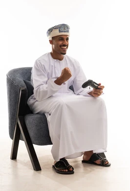 The fist is clenched tightly, waving the victory sign and achieving success. An Arab Gulf Omani man wearing a white dishdasha and a turban holds a game controller for a video game, with gestures of happiness and joy. Portrait with a white background, full-body.