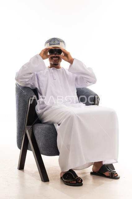 Looking at the camera using a telescope, an Arab Gulf Omani man wearing a white dishdasha and a turban sits on a chair and uses a telescope to magnify distant objects, full-body, portrait with a white background.
