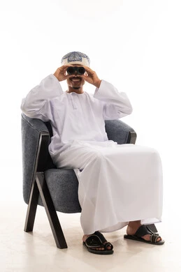 النظر الي الكاميرا باستخدام المنظار المقرب , رجل عربي خليجي عماني يرتدي دشداشة بيضاء و العمامة يجلس على الكرسي و يستخدم منظار لتقريب الاشياء البعيدة , الجسم بالطول الكامل , بورتريه خلفية بيضاء