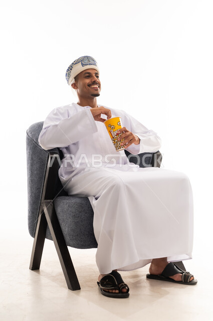 ايماءات الابتسامة و الاستمتاع اثناء مشاهدة الفيلم الكوميدي , رجل عربي خليجي عماني يرتدي دشداشة بيضاء و عمامة يأكل الفشار , الجسم بالطول الكامل , بورتريه خلفية بيضاء
