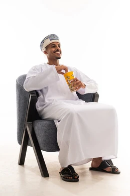 ايماءات الابتسامة و الاستمتاع اثناء مشاهدة الفيلم الكوميدي , رجل عربي خليجي عماني يرتدي دشداشة بيضاء و عمامة يأكل الفشار , الجسم بالطول الكامل , بورتريه خلفية بيضاء