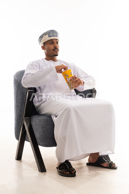 تعابير الاثارة و التشويق عند مشاهدة الافلام , رجل عربي خليجي عماني يرتدي دشداشة بيضاء و عمامة يجلس على كرسي رمادي اللون و يأكل الفشار , الجسم بالطول الكامل , خلفيةبيضاء