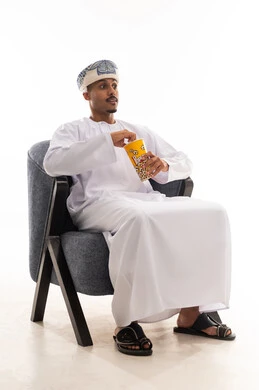 Expressions of excitement and suspense while watching movies, an Arab Gulf Omani man wearing a white dishdasha and a turban sitting on a gray chair and eating popcorn, full-body shot, white background.