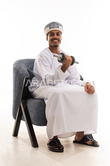 استعراض القوة البدنية امام الكاميرا , رجل عربي خليجي عماني يرتدي العمامة ودشداشة بيضاء يرفع الوزن الثقيل بثبات , خلفية بيضاء , الجسم بالطول الكامل , نظرة أمامية
