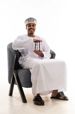 Expressions of happiness and joy after the passage of time, an Omani Arab man wearing a white dishdasha and turban sitting on a chair, holding the hourglass with delight, full-body, portrait with a white background.