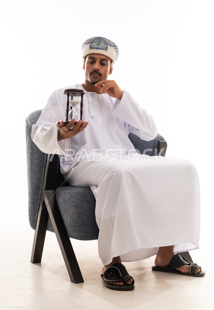 متابعة مرور الوقت ببطئ , رجل عربي خليجي عماني يرتدي الزي التقليدي في سلطنة عمان , النظر الي الساعة الرملية , الجسم بالطول الكامل , بورتريه خلفية بيضاء