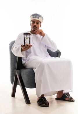 Watching time pass slowly, an Arab Gulf Omani man wearing traditional attire in the Sultanate of Oman, looking at the hourglass, full-length body, portrait with a white background.