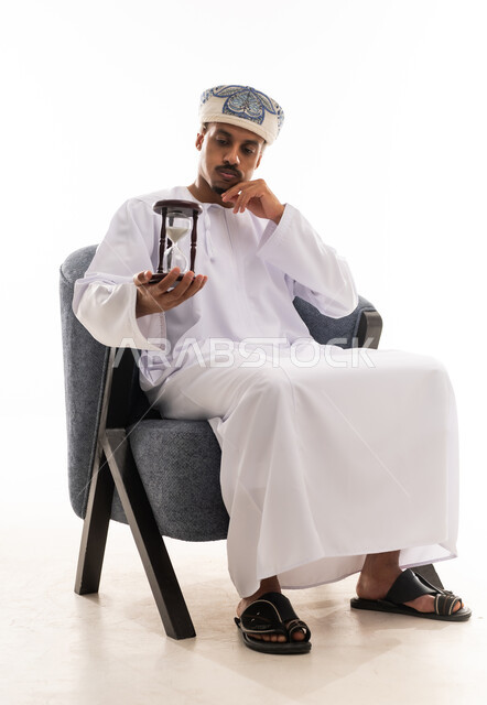 تعابير الملل اثناء الانتظار , رجل عربي خليجي عماني يرتدي دشداشة بيضاء و عمامة يحمل الساعة الرملية و يتابع الوقت , بورتريه خلفية بيضاء , الجسم بالطول الكامل