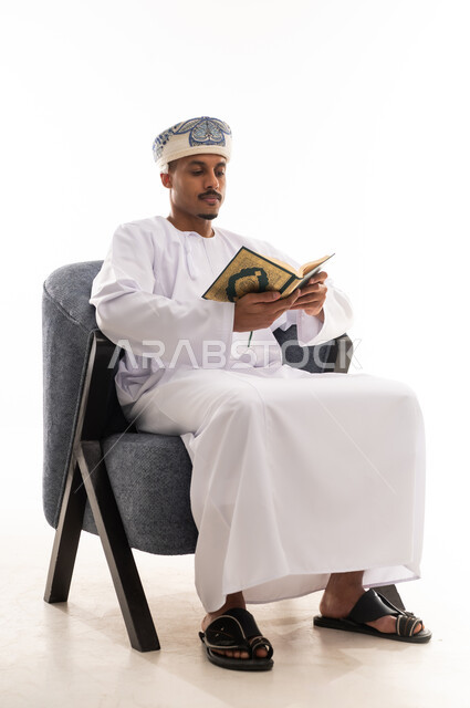 الشعور بالسكينة في الخلوة مع الله , رجل عربي خليجي عماني يرتدي دشداشة بيضاء و عمامة يحمل المصحف الشريف و يقرأ بامعان و تركيز , الجسم بالطول الكامل , بورتريه خلفية بيضاء