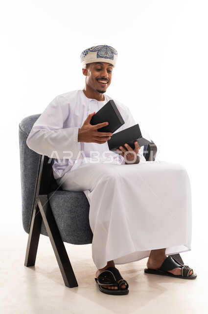 تعابير المفاجأة و الدهشة عند فتح الهدية , رجل عربي خليجي عماني يرتدي دشداشة بيضاء و عمامة يجلس على كرسي رمادي اللون , اياماءات السعادة و الفرحة , الجسم بالطول الكامل , بورتريه خلفية بيضاء