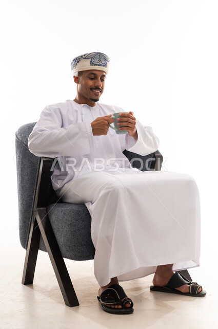 النظر الي الكوب الساخن و ابتسامة الاستمتاع بالمذاق الشهي , رجل عربي خليجي عماني يرتدي دشداشة بيضاء و العمامة يحمل القهوة العربية اللذيذة , بورتريه خلفية بيضاء , الجسم بالطول الكامل