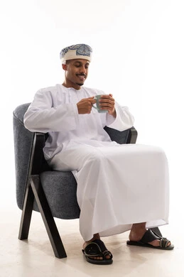 Looking at the hot cup and the smile of enjoyment from the delicious taste, an Arab Gulf Omani man wearing a white dishdasha and a turban holds the delicious Arabic coffee, portrait with a white background, full body.