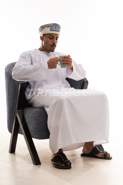تذوق النكهة المميزة للمشروب الساخن , رجل عربي خليجي عماني يرتدي دشداشة بيضاء و العمامة يحمل كوب دافئ , بورتريه خلفية بيضاء , الجسم بالطول الكامل